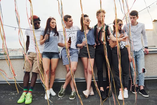 Leaning on the edge of the rooftop with decorates. Group of young people in casual clothes have a party together at daytime.