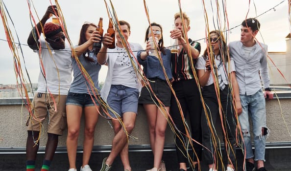 Leaning on the edge of the rooftop with decorates. Group of young people in casual clothes have a party together at daytime.