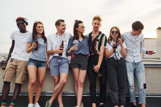 Standing with alcohol at the edge of rooftop. Group of young people in casual clothes have a party together at daytime.