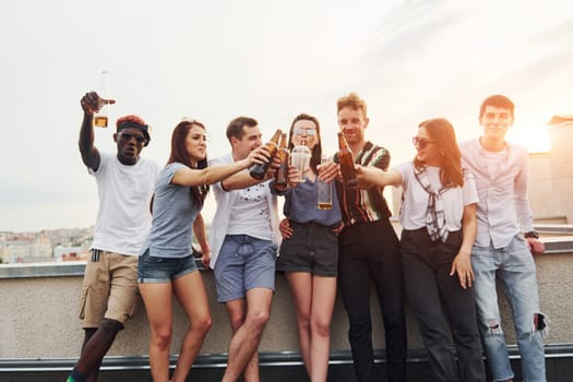 Standing with alcohol at the edge of rooftop. Group of young people in casual clothes have a party together at daytime.