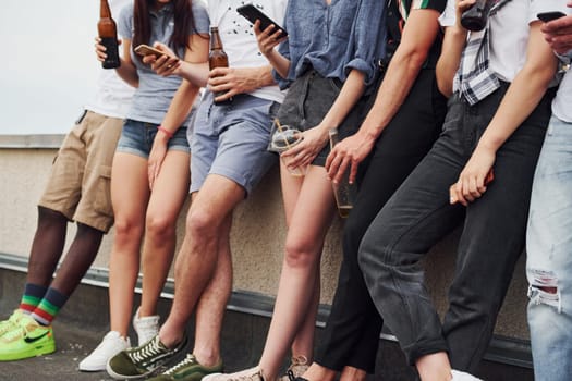 Standing with phones and alcohol in hands. Group of young people in casual clothes have a party at rooftop together at daytime.