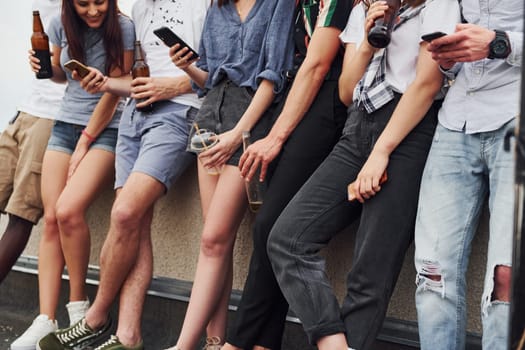 Standing with phones and alcohol in hands. Group of young people in casual clothes have a party at rooftop together at daytime.