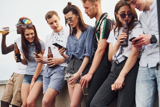 Standing with phones and alcohol in hands. Group of young people in casual clothes have a party at rooftop together at daytime.