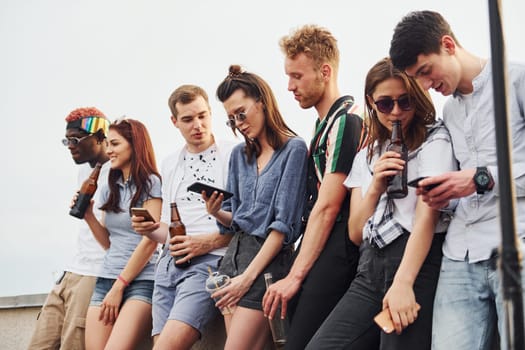 Standing with phones and alcohol in hands. Group of young people in casual clothes have a party at rooftop together at daytime.