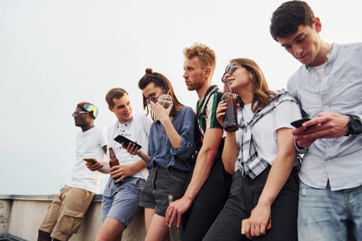 Standing with phones and alcohol in hands. Group of young people in casual clothes have a party at rooftop together at daytime.