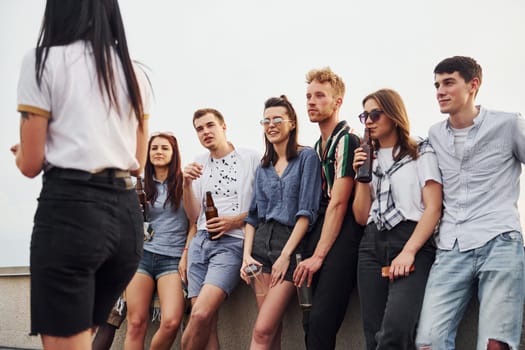 Standing with phones and alcohol in hands. Group of young people in casual clothes have a party at rooftop together at daytime.