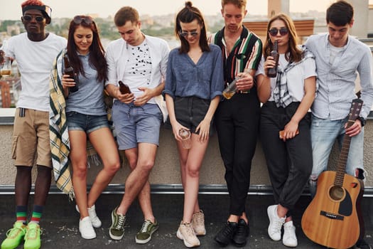 Standing with phones and alcohol in hands. Group of young people in casual clothes have a party at rooftop together at daytime.