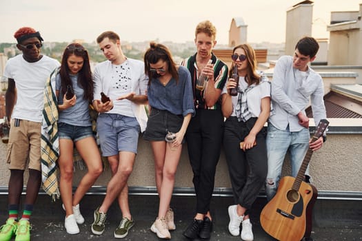 Standing with phones and alcohol in hands. Group of young people in casual clothes have a party at rooftop together at daytime.