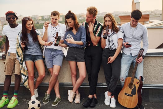 Standing with phones and alcohol in hands. Group of young people in casual clothes have a party at rooftop together at daytime.