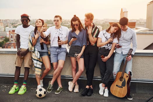 Standing with phones and alcohol in hands. Group of young people in casual clothes have a party at rooftop together at daytime.