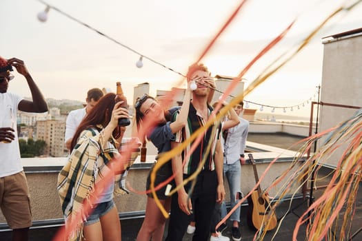 Playing game. Man's eyes covered by hands. Group of young people in casual clothes have a party at rooftop together at daytime.