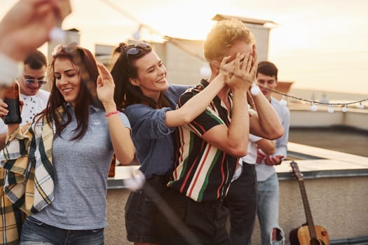 Playing game. Man's eyes covered by hands. Group of young people in casual clothes have a party at rooftop together at daytime.