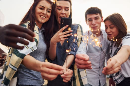 With sparklers in hands. Group of young people in casual clothes have a party at rooftop together at daytime.