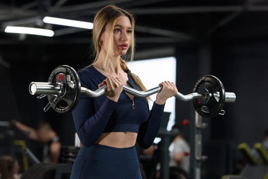 A young woman does exercises for biceps, a barbell with a curved bar. The woman in the gym. wearing blue leggins and long sleeve top.