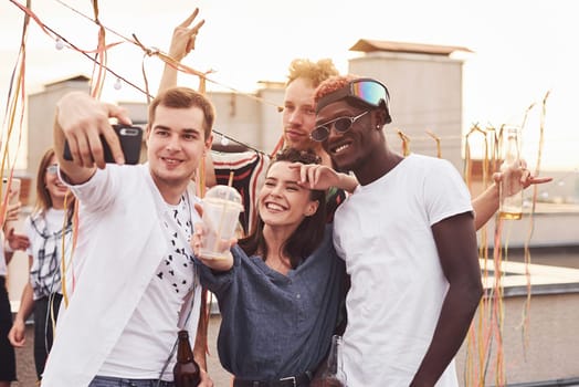 Standing together and taking selfie. Group of young people in casual clothes have a party at rooftop together at daytime.