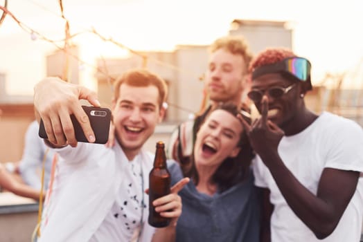 Standing together and taking selfie. Group of young people in casual clothes have a party at rooftop together at daytime.