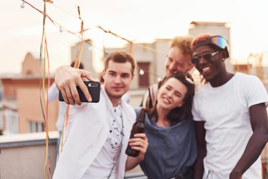 Standing together and taking selfie. Group of young people in casual clothes have a party at rooftop together at daytime.