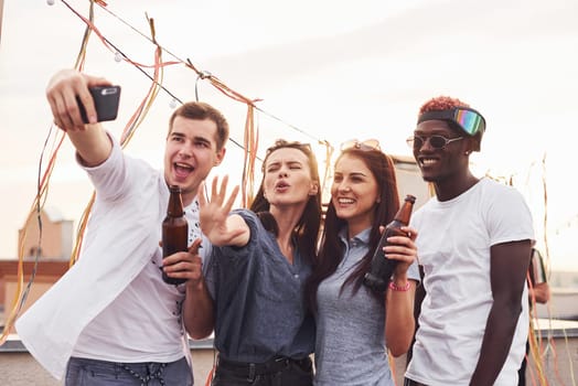 Standing together and taking selfie. Group of young people in casual clothes have a party at rooftop together at daytime.