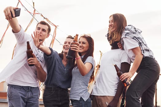 Standing together and taking selfie. Group of young people in casual clothes have a party at rooftop together at daytime.