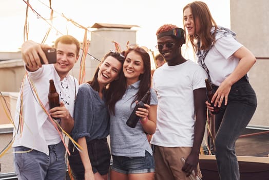 Standing together and taking selfie. Group of young people in casual clothes have a party at rooftop together at daytime.