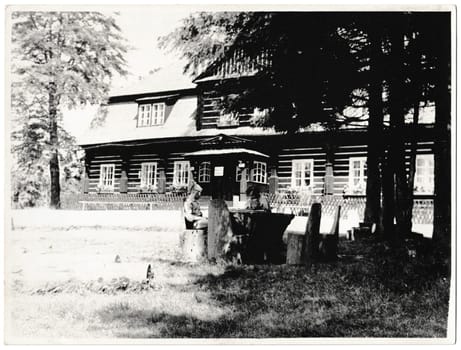 THE CZECHOSLOVAK SOCIALIST REPUBLIC - CIRCA 1960s: Retro photo shows wooden log cabin on the background. Two people sit in front of wooden cottage. Black & white vintage photography