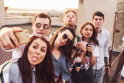 Making selfie by phone. Group of young people in casual clothes have a party at rooftop together at daytime.