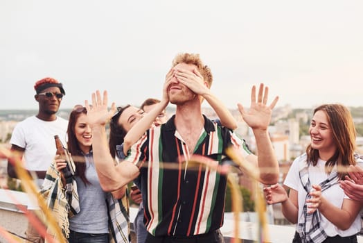 Man's eyes covered by hands. Group of young people in casual clothes have a party at rooftop together at daytime.