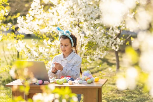 teenage girl paints Easter eggs with a laptop in the garden.