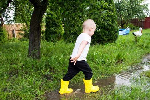 A small, bald-headed boy in yellow boots runs in the countryside through puddles in the fresh air