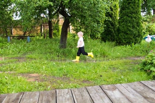 A small, bald-headed boy in yellow boots runs in the countryside through puddles in the fresh air