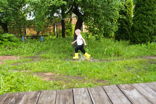 A small, bald-headed boy in yellow boots runs in the countryside through puddles in the fresh air