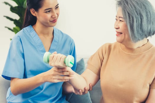 Caregiver helping contented senior woman exercise with dumbbell at home. Rehab for elder, senior medical care.