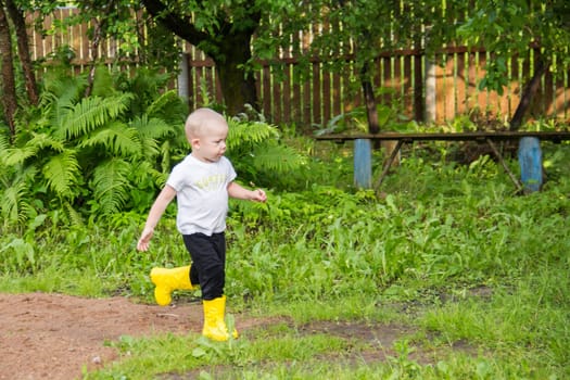 A small, bald-headed boy in yellow boots runs in the countryside through puddles in the fresh air