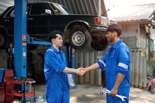 Two Asian automotive mechanic repairman handshake in garage. Vehicle service manager working in mechanics workshop, feel happy and success after check and maintenance car engine for customer..
