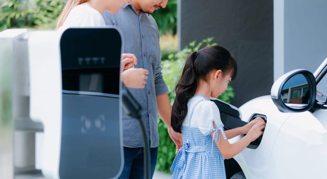 Progressive young parents and daughter with electric vehicle and home charging station. Green and clean energy from electric vehicles for healthy environment. Eco power from renewable source at home.