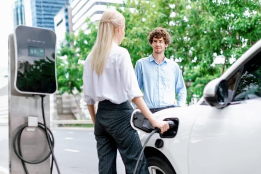 Progressive businessman and businesswoman with electric car parking and connected to public charging station before driving around city center. Eco friendly rechargeable car powered by clean energy.