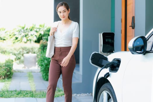 Progressive asian woman and electric car with home charging station. Concept of the use of electric vehicles in a progressive lifestyle contributes to a clean and healthy environment.