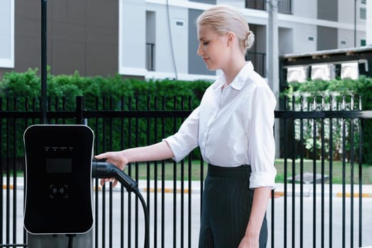 Progressive businesswoman insert charger plug from charging station to her electric vehicle with apartment condo building in background. Eco friendly rechargeable car powered by sustainable energy.