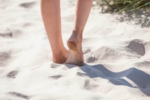 Naked legs of a woman walking on the white sand of the sea on a summer day