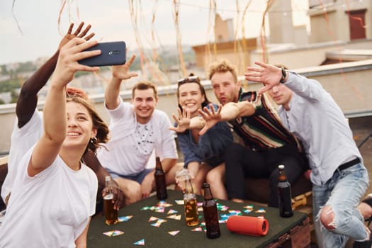 Girl doing photo when people playing card game. Group of young people in casual clothes have a party at rooftop together at daytime.