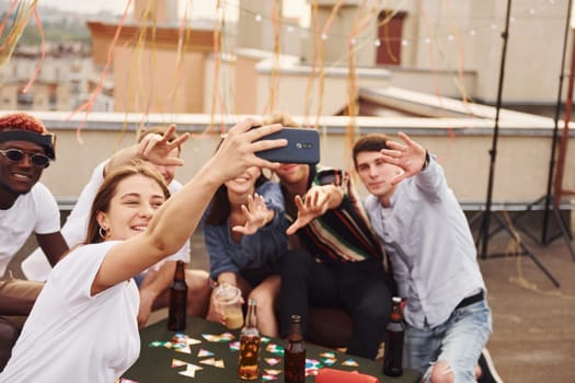 Girl doing photo when people playing card game. Group of young people in casual clothes have a party at rooftop together at daytime.