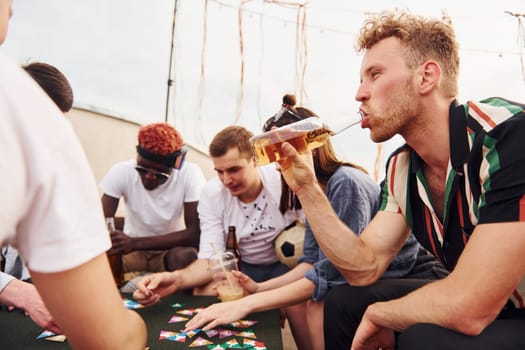 Playing card game. Group of young people in casual clothes have a party at rooftop together at daytime.