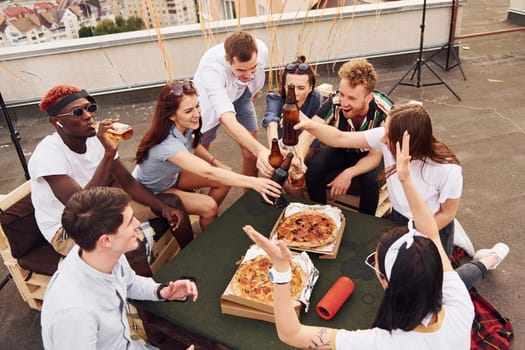 Doing cheers by bottles with beer. Group of young people in casual clothes have a party at rooftop together at daytime.