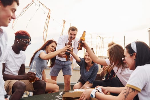 Doing cheers by bottles with beer. Group of young people in casual clothes have a party at rooftop together at daytime.