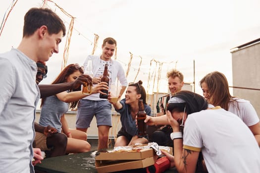 Doing cheers by bottles with beer. Group of young people in casual clothes have a party at rooftop together at daytime.