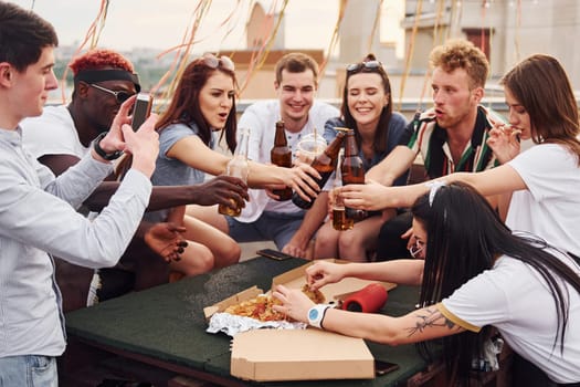 Doing cheers by bottles with beer. Group of young people in casual clothes have a party at rooftop together at daytime.