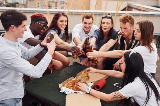 Doing cheers by bottles with beer. Group of young people in casual clothes have a party at rooftop together at daytime.