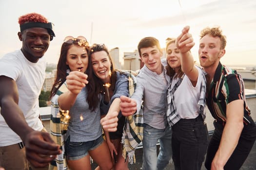 With sparklers in hands. Group of young people in casual clothes have a party at rooftop together at daytime.