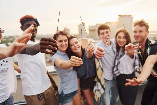 With sparklers in hands. Group of young people in casual clothes have a party at rooftop together at daytime.