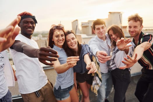 With sparklers in hands. Group of young people in casual clothes have a party at rooftop together at daytime.